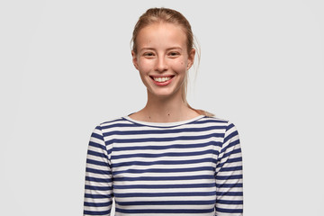 Cute female teenager has broad smile, wears striped sweater, being in good mood after unforgettable...