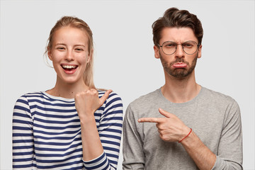 Happy girlfriend makes jokes of her boyfriend, indicates with thumb at him and laughs joyfully....