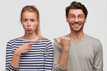 Displeased abused female frowns face as blames her husband of something, indicates with fore...
