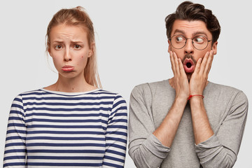 Upset female model with sullen expression being in low spirit after quarrel with boyfriend stands near him. Surprised young man keeps hands on face, wonders something. People and emotions concept