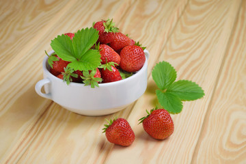 Two strawberries and a white bowl filled with strawberries on a light wooden table.