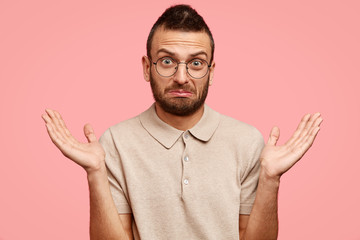 Studio shot of displeased bearded hipster male has clueless expression and puzzled look, doesn`t know from what to start work, has many tasks, not much time, isolated over pink studio background