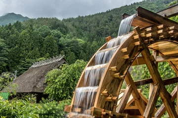 Foto op Plexiglas Molens Het molenwiel draait onder een stroom van water, achtergrond van dorp met traditionele huizen met rieten daken