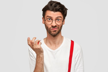 Fototapeta premium Frustrated puzzled male with European appearance, frowns face and raises hand, feels embarrassed after recieving difficult task at work, sees something not clear, isolated over white studio wall