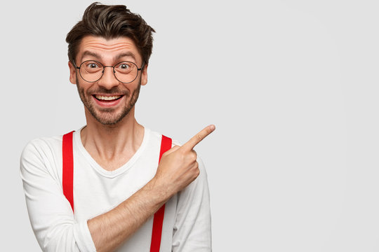 Happy excited male model shows blank space against white background, has dark stubble, trendy haircut, rejoices selling new items in shop, has great mood, expresses optimism and positiveness