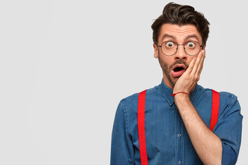 Portrait of excited hipster male with stubble, keeps hand on cheek, wears denim shirt with red braces, stares at camera in surprisement, recives unexpected news, poses against white wall, blank space