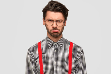 Young fashionable male has stubble, gloomy expression, being dissatisfied with results of work, dressed in stylish outfit, wears round glasses, frowns face in displeasure. Negative facial expressions