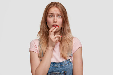 Cute frightened European woman looks with puzzled expression, recieves bad news from relatives, has wide opened eyes in stupor, poses against white studio background. People and reaction concept