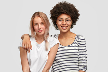 Two female mutiethnic lesbians embrace each other and smile joyfully, pose at camera with happy expressions, isolated over white background. African American woman hugs her European companion