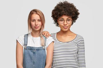 Multiethnic women of different races, have confident and pleased expressions at camera, stand close to each other. Pretty Caucasian female in denim overalls meets with her best African friend