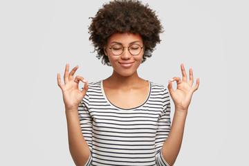 Horizontal shot of happy dark skinned female makes okay gesture, keeps eyes closed from pleasure, demostrates her agreement, has positve expression. Cheerful mixed race young woman poses indoor