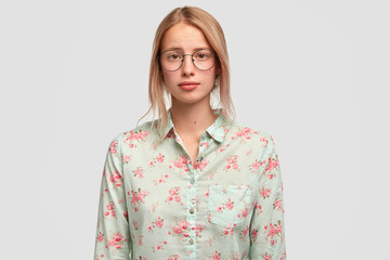 Waist up portrait of beautiful European female with pleading expression, has clean skin, wears big glasses and flower print blouse, stands against white background. Lovely student rests after classes