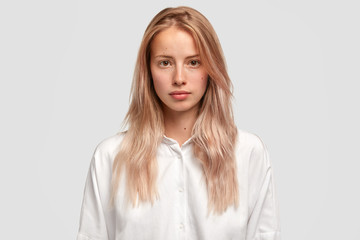 Headshot of self assured Caucasian female with healthy pure skin, wears loose white shirt, looks directly into camera, has serious expression, contemplates about future plans. Beauty and lifestyle