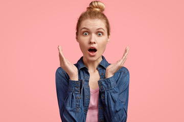 People, emotions and facial expressions concept. Stupefied beautiful blonde female youngster gestures with hands and stares at camera, dressed in denim jacket, stands against blank pink wall