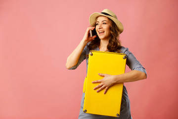 Young happy beautiful woman in hat holding yellow suitcase and talking by mobile over pink background