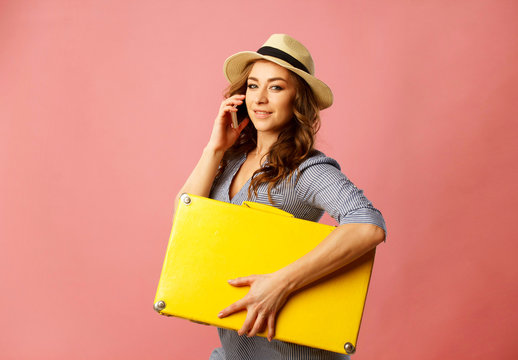 Young happy beautiful woman in hat holding yellow suitcase and talking by mobile over pink background