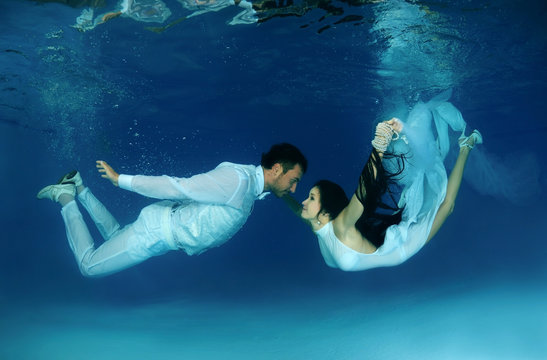Bride and groom in a white wedding dress swim to each other underwater in the pool. Underwater wedding