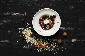 Cereal served with blueberries, blackberries, raspberries, cherries and whipped cream in plate standing on blue old wooden table with ground oats, walnuts and date fruits scattered around. Top view.