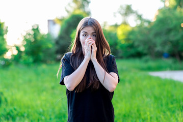 A brunette girl with long summer hair in nature in black T-shirt. Hands covering his mouth. Emotionally, fear is a surprise. Dressed in casual clothes. Large Surprised brown eyes.