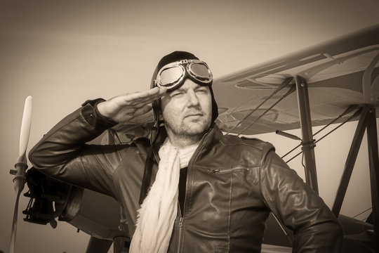
Portrait of a vintage pilot with leather cap, scarf and aviator glasses salutes in front of a historical biplane - Portrait of a man in historical pilot clothing - vintage old picture style