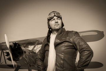 Portrait of a vintage pilot with leather cap, scarf and aviator glasses in front of a historic airplane biplane - Portrait of a man in historical pilot clothing - vintage old picture style
