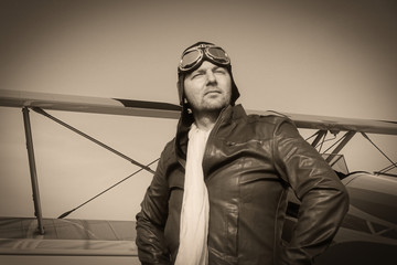 Portrait of a vintage pilot with leather cap, scarf and aviator glasses in front of a historic airplane biplane - Portrait of a man in historical pilot clothing - vintage old picture style
