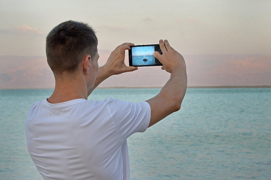 Guy Tourist in a white shirt takes pictures using a smartphone stone in the sea at sunset. The Dead Sea, Israel. Dreamy mountain landscape of Jordan, Rear view. Concept Technology and smartphones