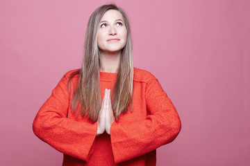 Young female with long straight hair looking up hopefully praying with her hands folded, hoping for best before important event in her life. Lovely European lady in red begging for something good.