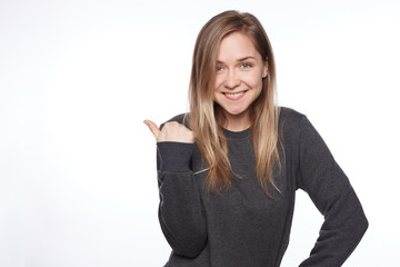 Obraz premium Charming long blond haired teenage lady shows copy space. Pleased lovely woman gives good advice, indicates with thumb, has happy expression. Glad tender female poses indoor over wall. Look there!