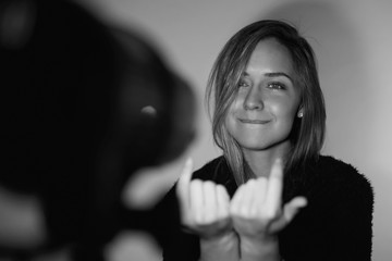 Attractive young female actress wearing black fur jacket looking tenderly at camera in closed posture, showing come on gesture, standing against white wall during audition under light of soffits.