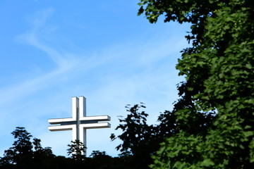 white big cross against the sky shining through green tree crowns.
