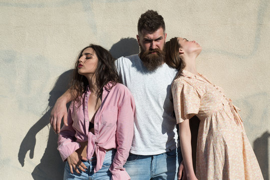 Man with beard hugs two ladies on hot sunny day. Girls turned to opposite sides while man hugs them. Love triangle concept. Threesome suffers of heat on summer day, wall on background