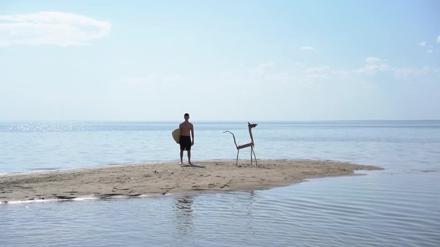 A young man with a surfboard is standing by a wooden animal made of sticks on a sandy island in the middle of the water. Abstract summer video concept.
