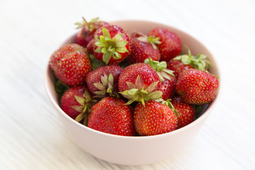 Fresh strawberries in a pink ceramic bowl on white wooden table, side view.