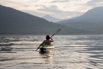 Woman kayaking around the beautiful Canadian Mountain Landscape during a vibrant cloudy evening. Taken in Howe Sound, North of Vancouver, British Columbia, Canada.