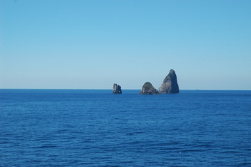 Three jagged rocks rising from a blue ocean. The sky is clear.