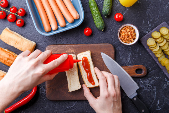 Picture on top of table with ingredients for hot dogs, cutting board, man's hands