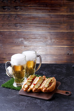 Photo of two mugs of foam beer, green grass with football, hotdogs