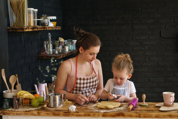 Young mother and daughter prepare cookies in kitchen. They are in aprons. Little girl is looking at baking dish. Family time.