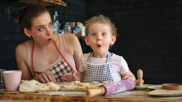 Young mother and daughter prepare cookies in kitchen. They are in aprons. Little girl and mother surprise and admiration. Positive emotions. Family time.