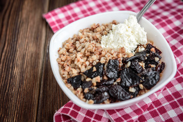 Boiled buckwheat with milk, prune and cottage cheese in white bowl on dark wooden table
