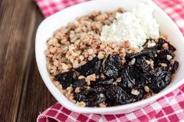 Boiled buckwheat with milk, prune and cottage cheese in white bowl on dark wooden table