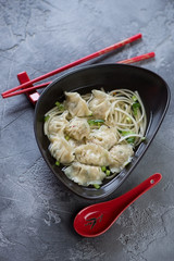 Black bowl of wonton noodle soup over grey concrete background, vertical shot, selective focus