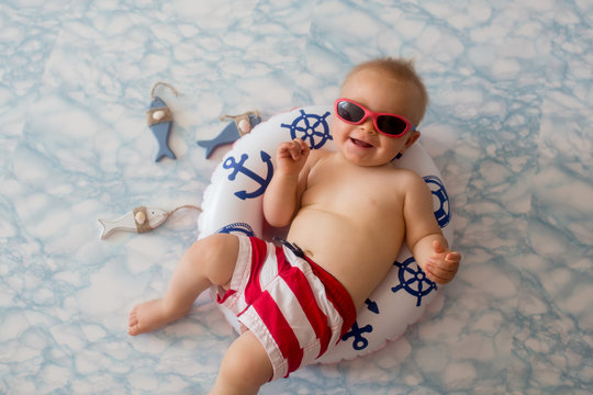 Cute baby boy lying down on a tiny inflatable swim ring,  wearing swimsuit shorts and sunglasses, indoor shot