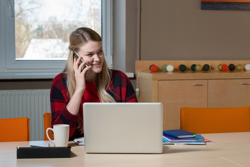 A blonde woman sitting at a laptop and talking over a cell phone.