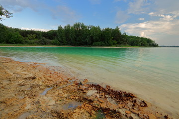 Brzeg sztucznego zbiornika wodnego nazywanego Lazurowym jeziorem, okolice Turku, Polska, wypełnionego chemicznymi odpadami z elektrowni, siarkowy osad na brzegu, w tle roślinność na przeciwnym brzegu,