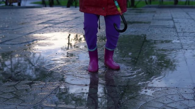 A cute little girl in a red raincoat jumping over a puddle in the park at sunset. The child smiles and enjoys the fun, slow motion.