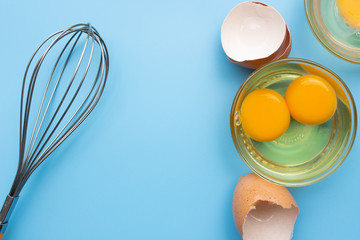 Fototapeta premium Raw fresh egg in glass bowl with eggshell and whisk on blue background top view with copy space