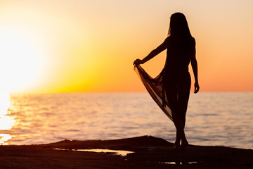 silhouette fo a pretty young woman with long dark hair in black transparent dress near the sea over sunset. concept of happy holiday and resort time.