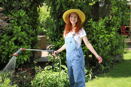 H&uuml;bsche rothaarige Frau w&auml;ssert lachend mit einer Handbrause ein Beet im Garten